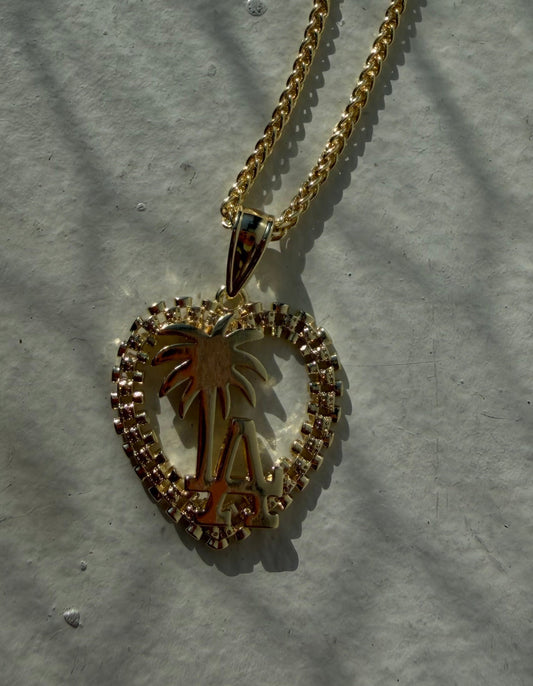 A gold heart-shaped pendant necklace featuring a palm tree design, resting on a light gray textured surface with soft shadows.