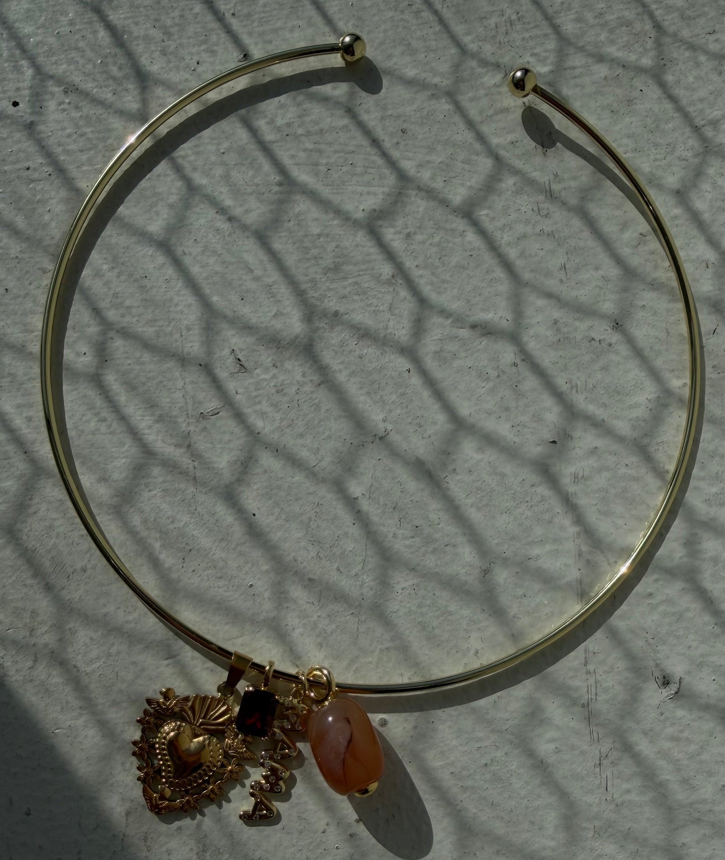 A thin gold-toned bangle bracelet with a dangling ornate gold charm and a small peach-colored bead, photographed on a light textured surface with strong shadows.