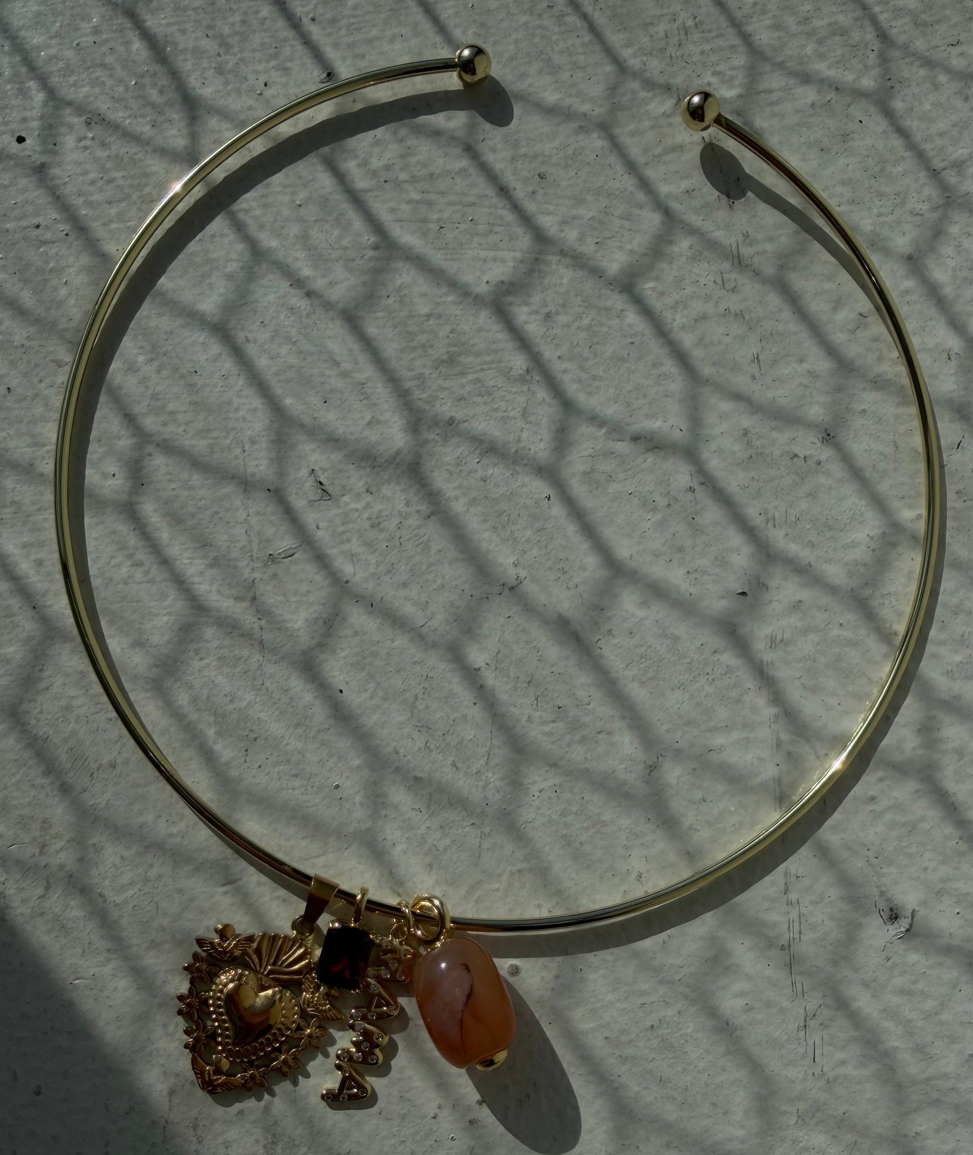 A thin gold-toned bangle bracelet with a dangling ornate gold charm and a small peach-colored bead, photographed on a light textured surface with strong shadows.