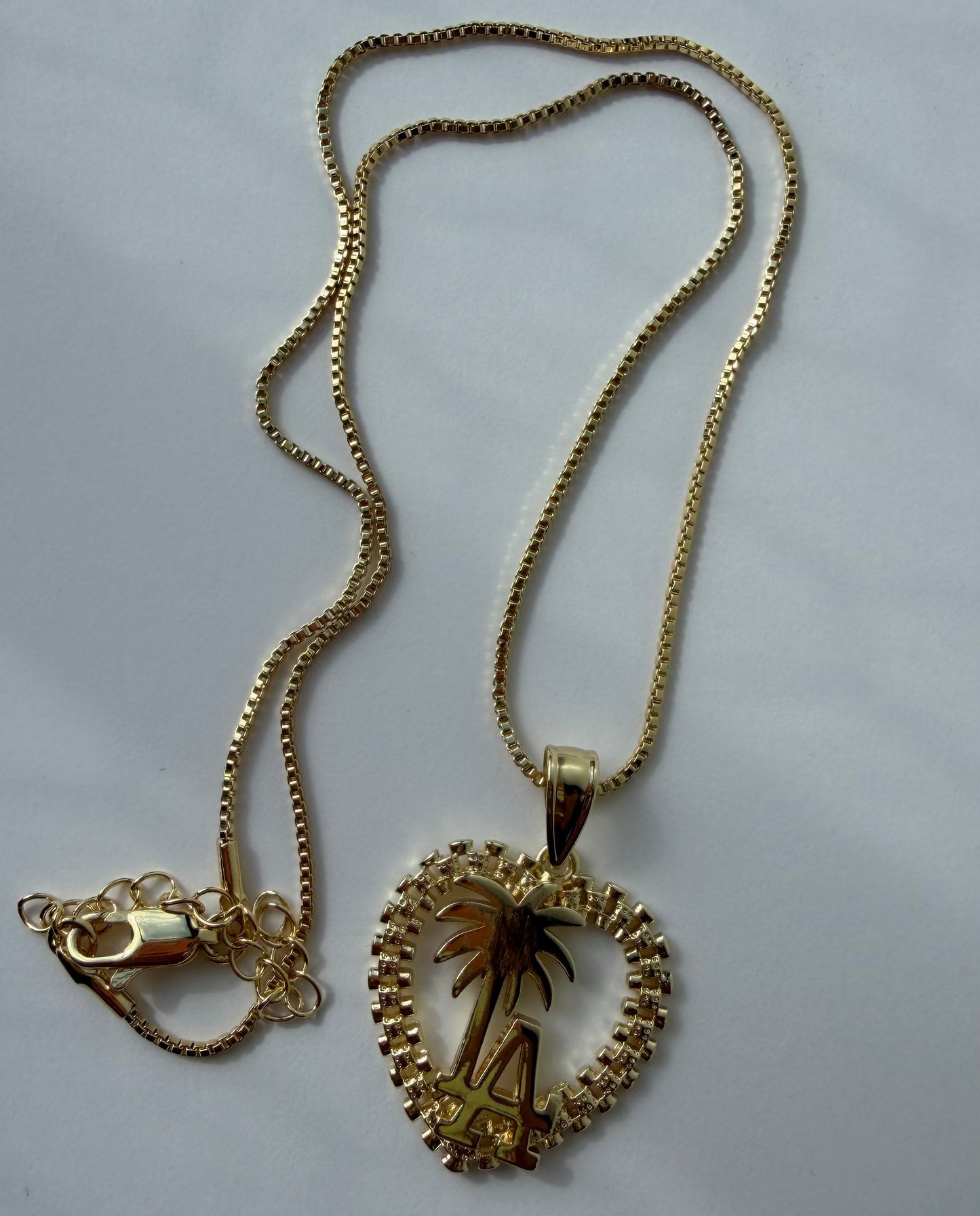 A gold chain necklace with a heart-shaped pendant featuring a palm tree design, displayed on a light gray background.