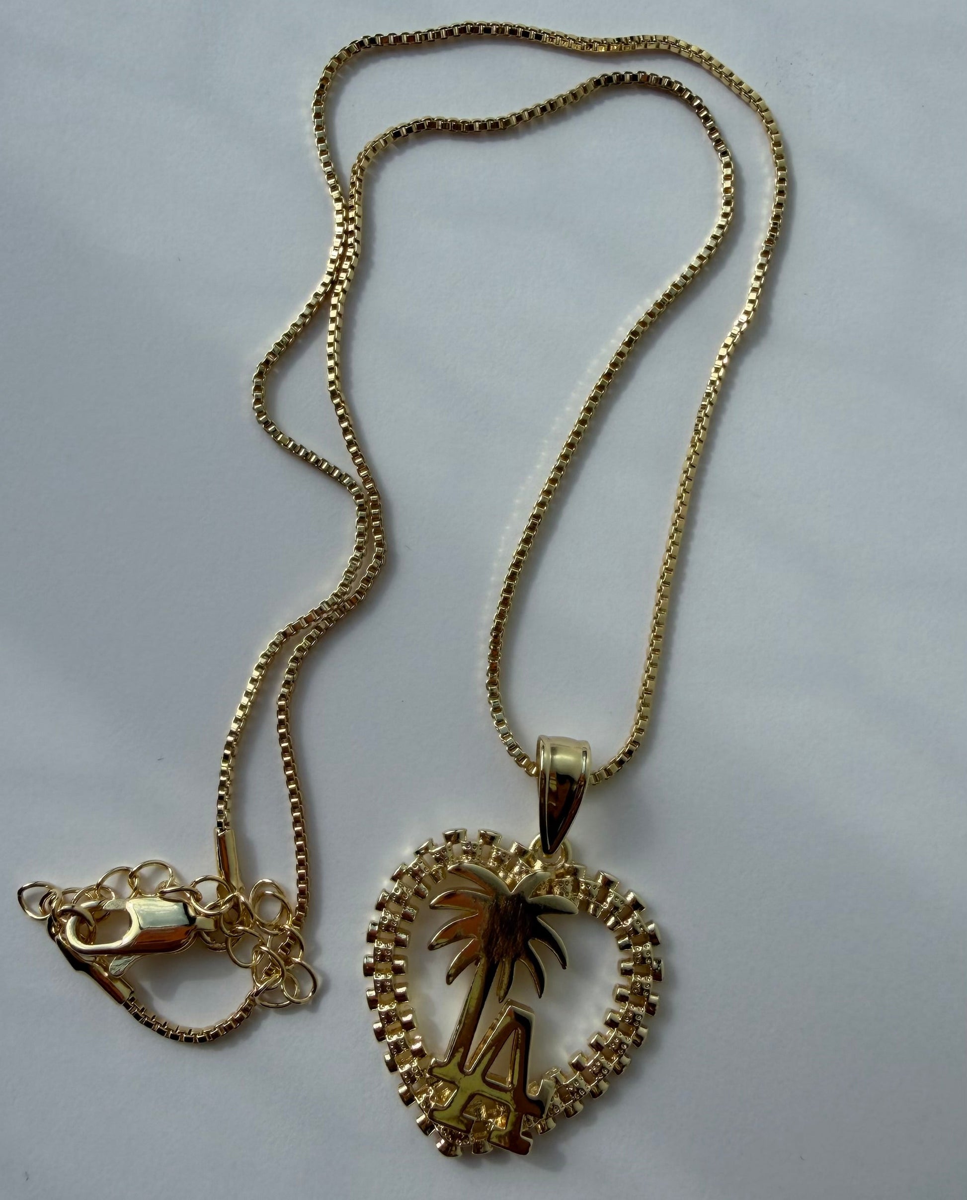A gold chain necklace with a heart-shaped pendant featuring a palm tree design, displayed on a light gray background.