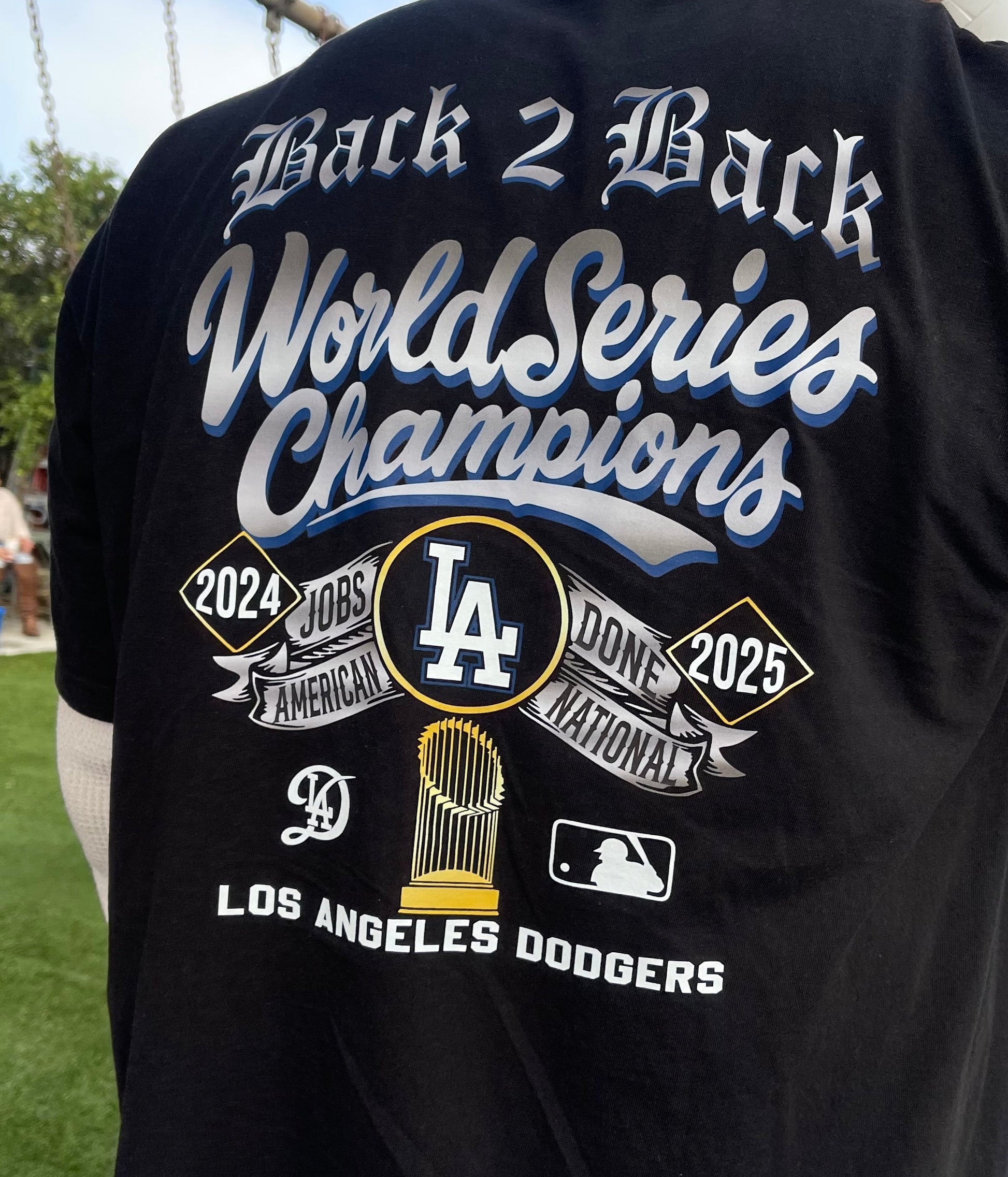 Black t-shirt with 'Back 2 Back World Series Champions' text and Los Angeles Dodgers logo on a grassy field.